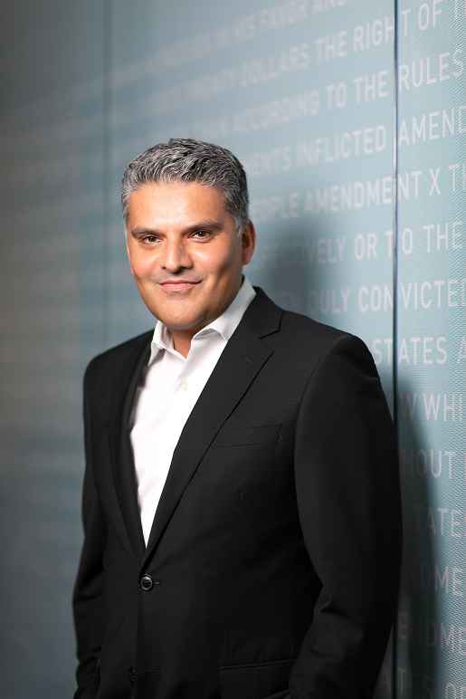 Chandra Bhatnagar (a brown skinned man with salt and pepper hair wearing a white dress suit and black suit jacket) standing in front of a light blue wall with white lettering.