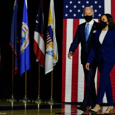 President-Elect Joe Biden and Vice President-Elect Kamala Harris arrive on stage at campaign event with American flag in background.