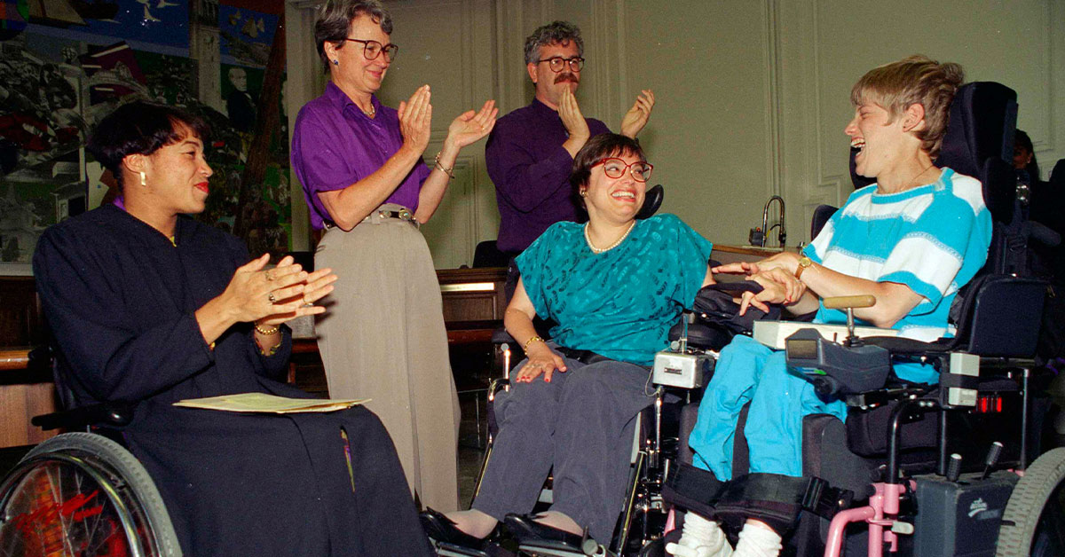 Judy Heumann, center wheelchair, was applauded as she was sworn in as U.S. Assistant Secretary for Special Education and Rehabilitative Service by Judge Gail Bereola, left, on June 29, 1993. Standing at left is Berkeley Mayor Loni Hancock with sign language interpreter Joseph Quinn, and Julie Weissman, right.