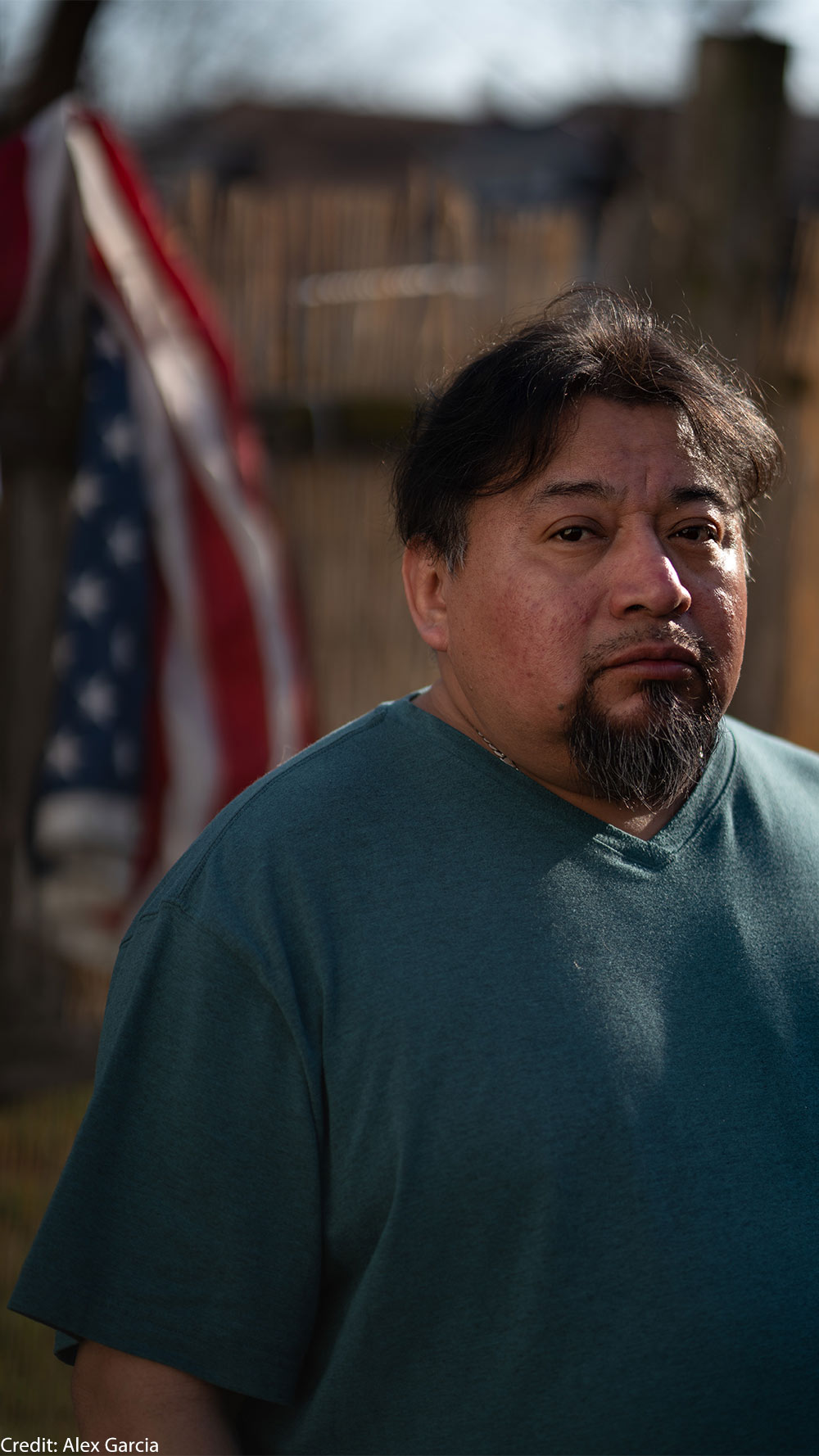 Margarito Casta​​ñon Nava looks into the camera as an American flag hangs from a wooden fence behind him.