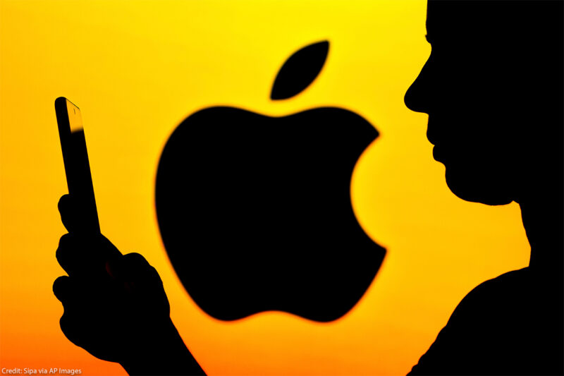In shadow, a person holds a phone with Apple logo (also in shadow) against a yellow background.