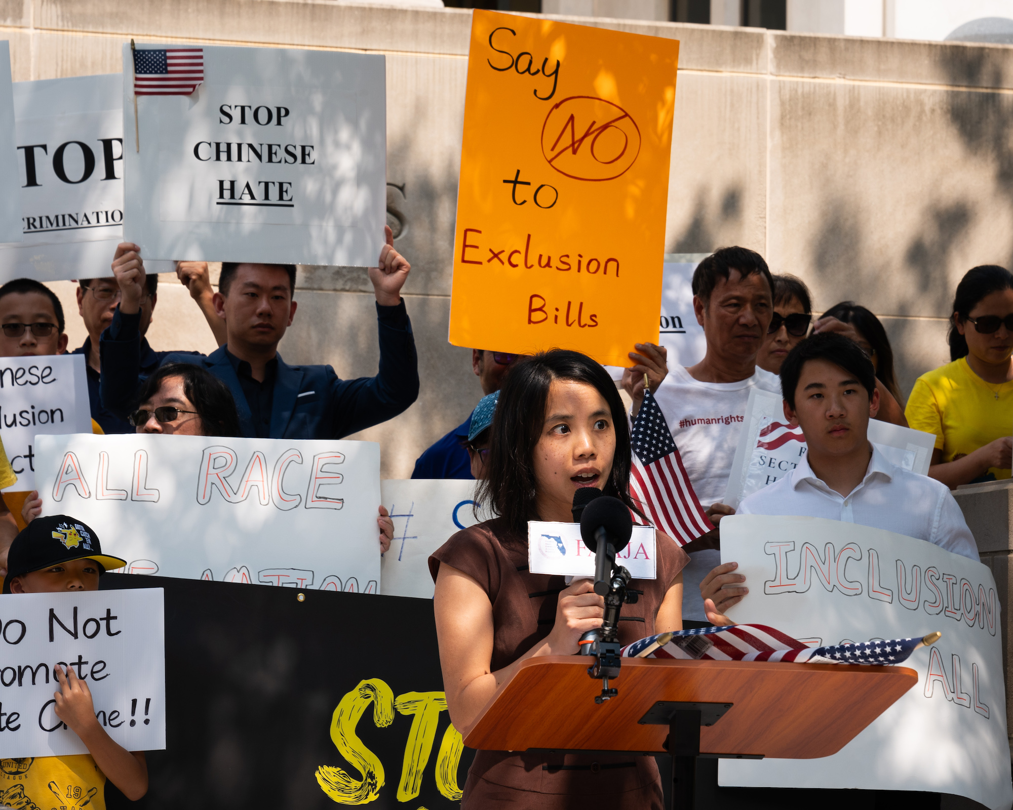 A woman speaks in front of a crowd with signs advocating against race-based violence in the name of national security.