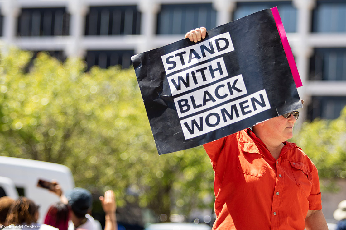 Someone holding a sign saying Stand With Black Women.