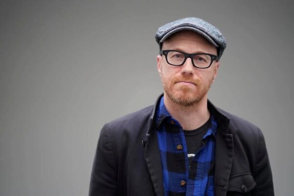 Kliph Nesteroff (a white man with a red short beard wearing black framed glasses, a gray newspaper boy hat, a blue and black flannel button up shirt and a navy blue jacket) stands in front of a gray background.