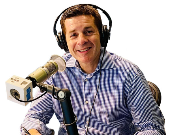 Dean Obeidallah (a pale skinned man wearing a checkered blue and white button up shirt and headphones) sits in front of a microphone.